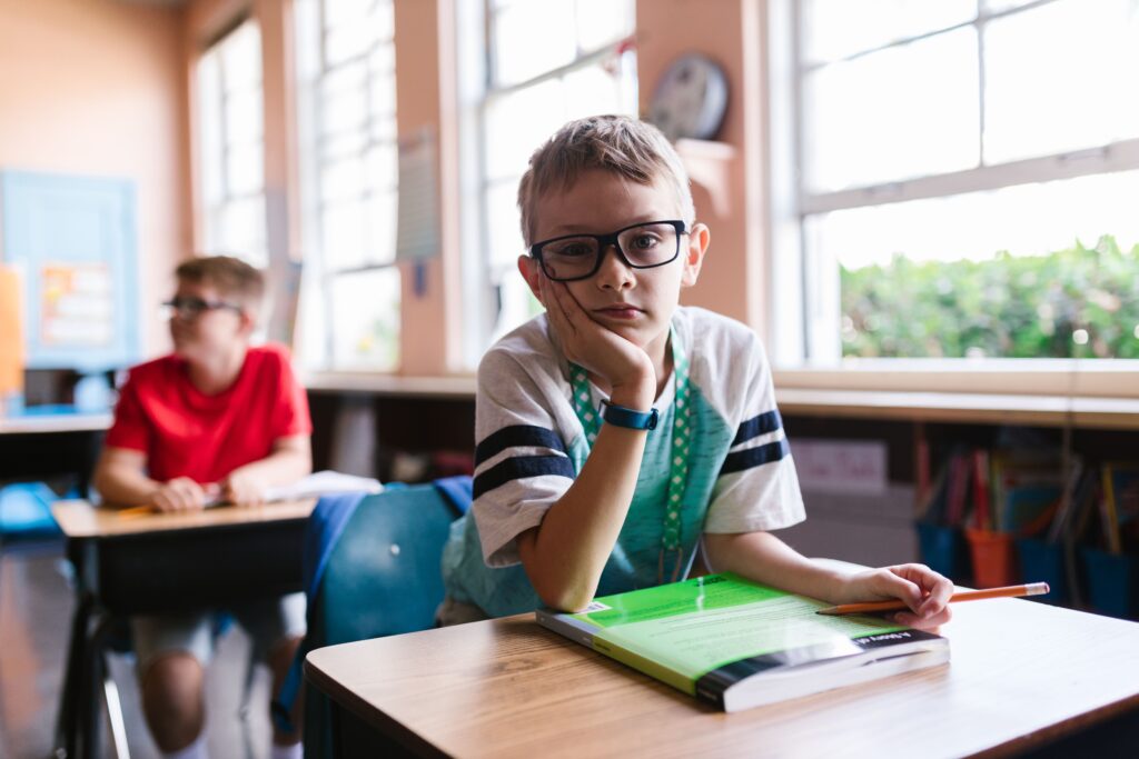 Young boy with glasses in a classroom, pen in hand, looking thoughtful.